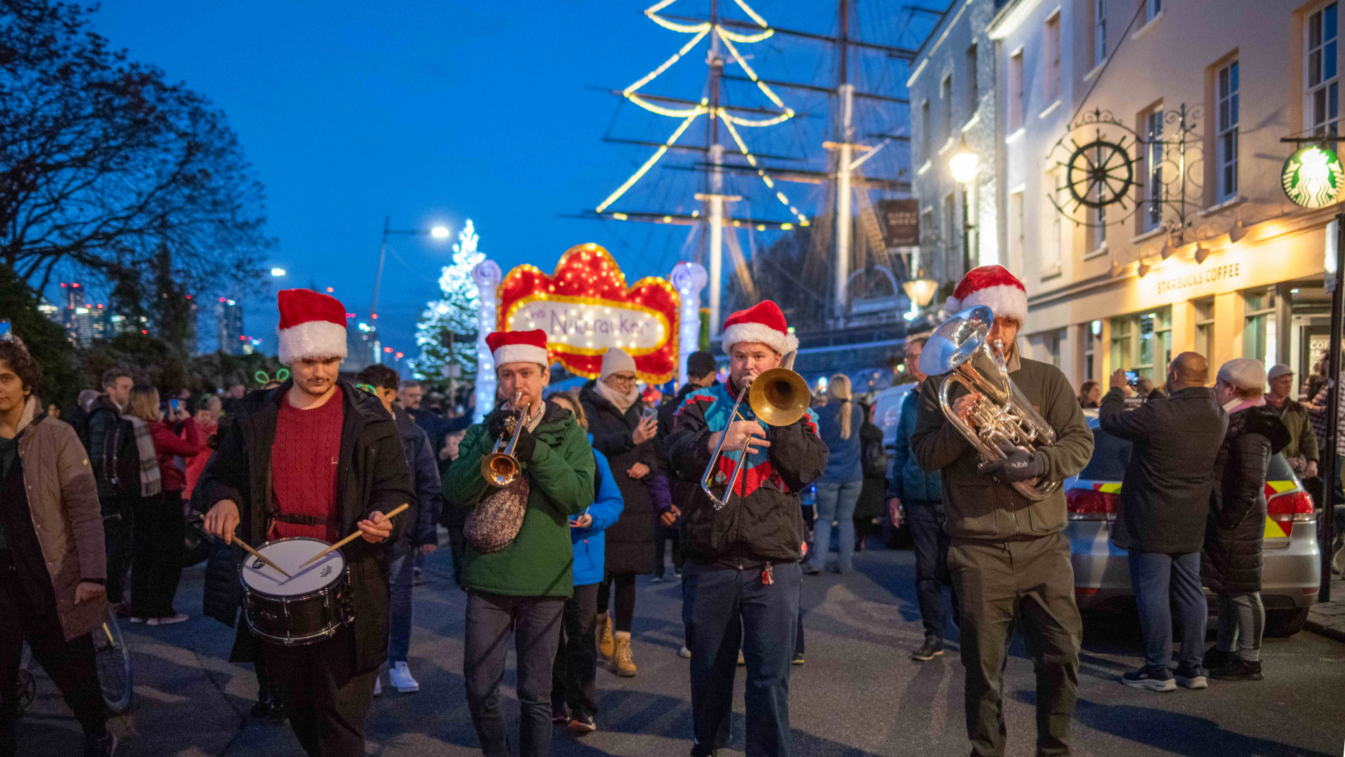 A four piece band walk down the street in front of Greenwich wearing Christmas hats surrounded by people holding lanterns and lights, with a view of Cutty Sark in the background decorated with lights in the shape of a Christmas tree