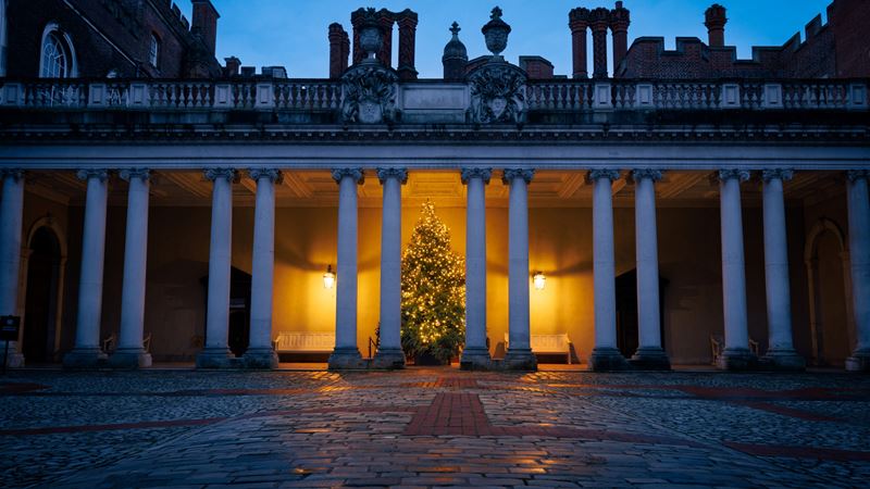 See the iconic Hampton Court Palace dressed up for the festive season in the style of the Tudors. Credit: Historic Royal Palaces. Image courtesy of Historic Royal Palaces. A photo of a Christmas tree glowing under one of the walkways at Hampton Court Palace at night with columns in the foreground
