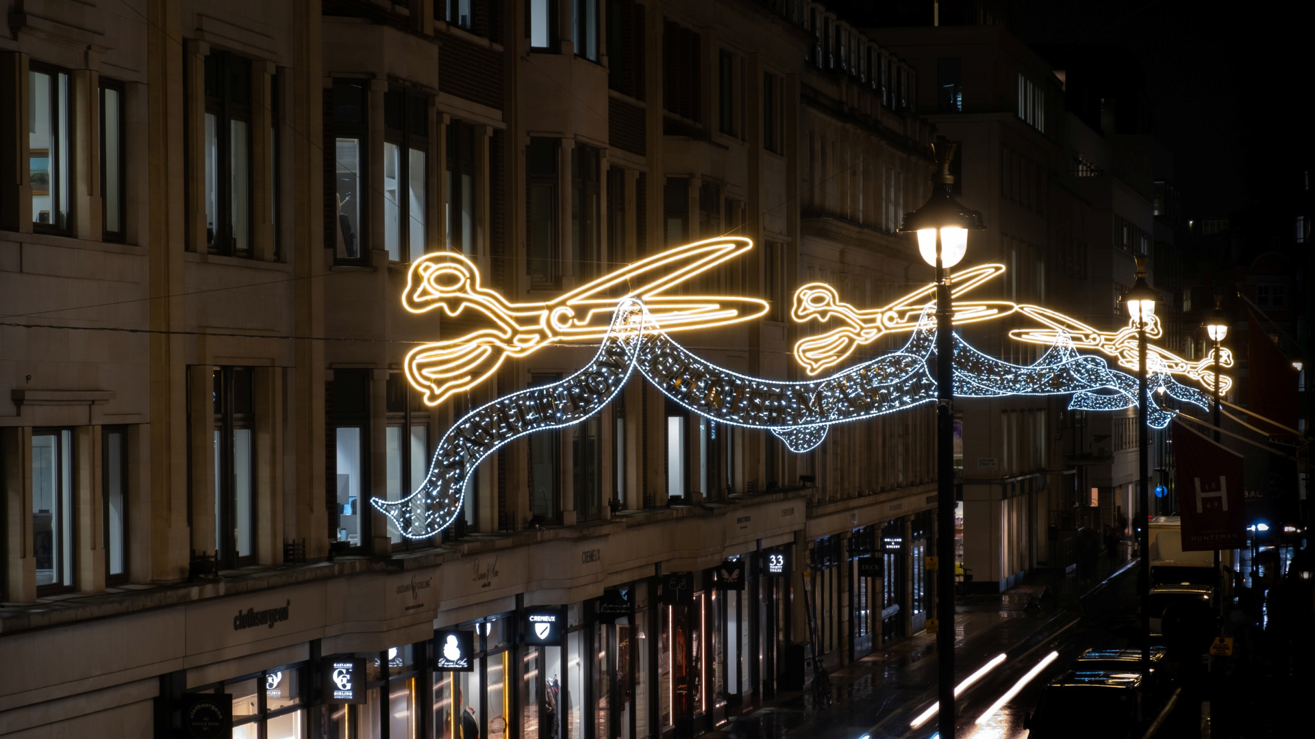 A photo of the famous Savile Row street at night with giant light up shears cutting fabric hanging up high for the festive season with street lamps and light trails from cars below