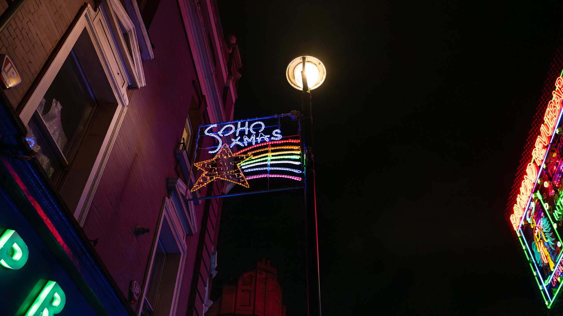 A rectangular panel of Christmas lights hang over Soho in the night sky with a shooting star in rainbow colours and the words 