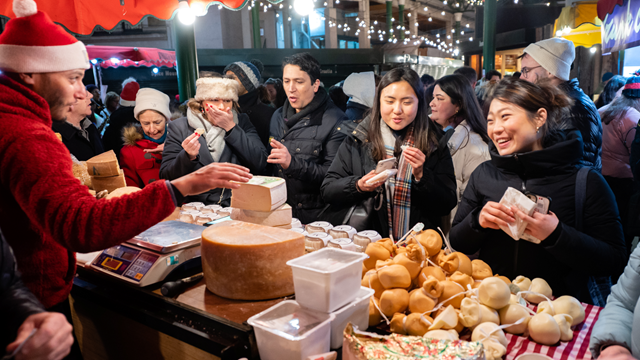 Crowds of people wrapped in warm clothes queue for a cheese stall at Borough Market stocked with piles of cheeses of different shapes, sizes and colours whilst a man behind a counter wearing a Christmas hat weighs cheese. 