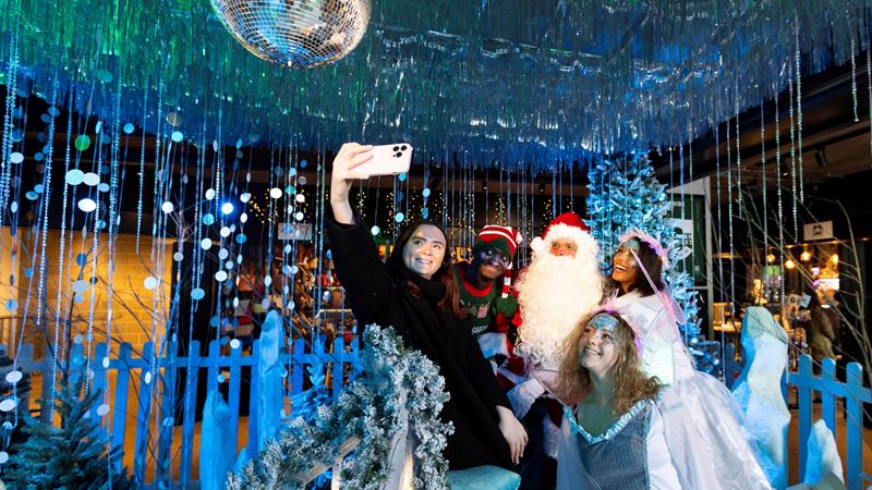 Explore Camden as it's turned into a magical festive wonderland with twinkling lights and an ice palace grotto! Credit: David Parry. Image courtesy of Sister London. A photo of four friends dressed as elves taking a selfie on a phone with Santa inside an ice palace themed grotto with shiny decorations and a disco ball