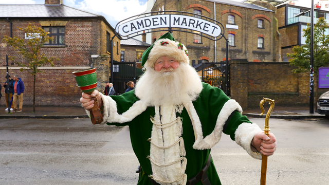A man dressed up as Santa in a green velvet costume and matching hat with a white beard ringing a bell outside of the giant Camden Market entrance sign