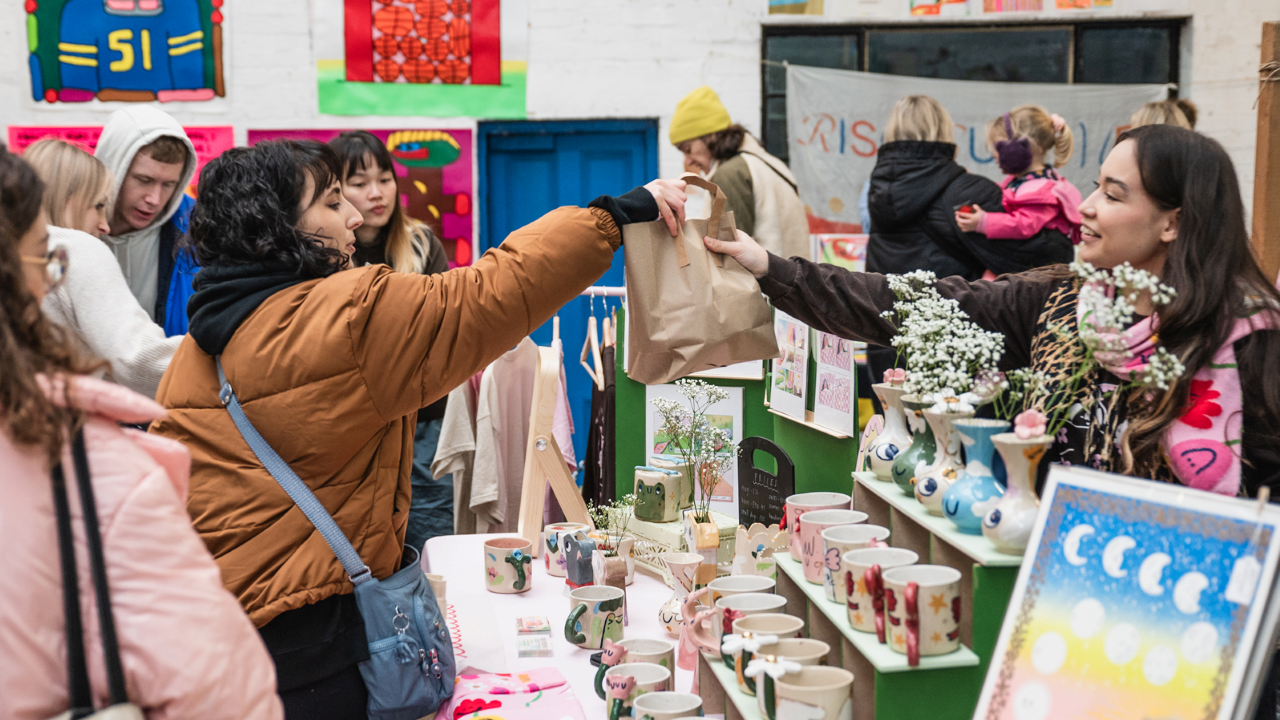 A young woman in a brown puffer jacket with a blue shoulder bag takes a brown paper bag filled with goodies from an independent seller at a market, with her stall full of goodies like colourful handmade mugs and painted canvases
