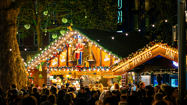 Warme strahlende Lichter erhellen eine Holzhütte im Leicester Square Weihnachtsmarkt, ein Spielzeugsoldat befindet sich unter dem Dach und Menschenmengen befinden sich vor der Hütte.