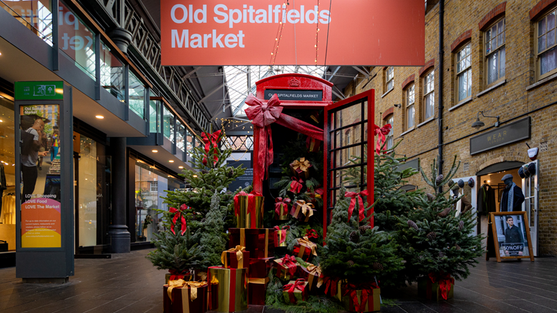 Find unique stocking fillers at Old Spitalfields Christmas market. Credit: Old Spitalfields Market. Image courtesy of Old Spitalfields Market. A red phone box stands under an Old Spitalfields Market sign with the door open and presents spilling out, surrounded by Christmas trees