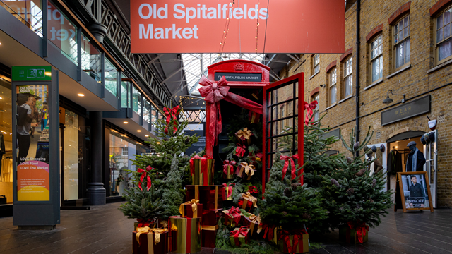 A red phone box stands under an Old Spitalfields Market sign with the door open and presents spilling out, surrounded by Christmas trees