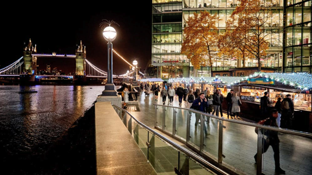 Personen laufen am Abend an Weihnachtsverkaufsständen vorbei, die entlang der Themse stehen und mit LED Lichtern geschmückt sind, die Tower Bridge ist in der Ferne zu sehen.