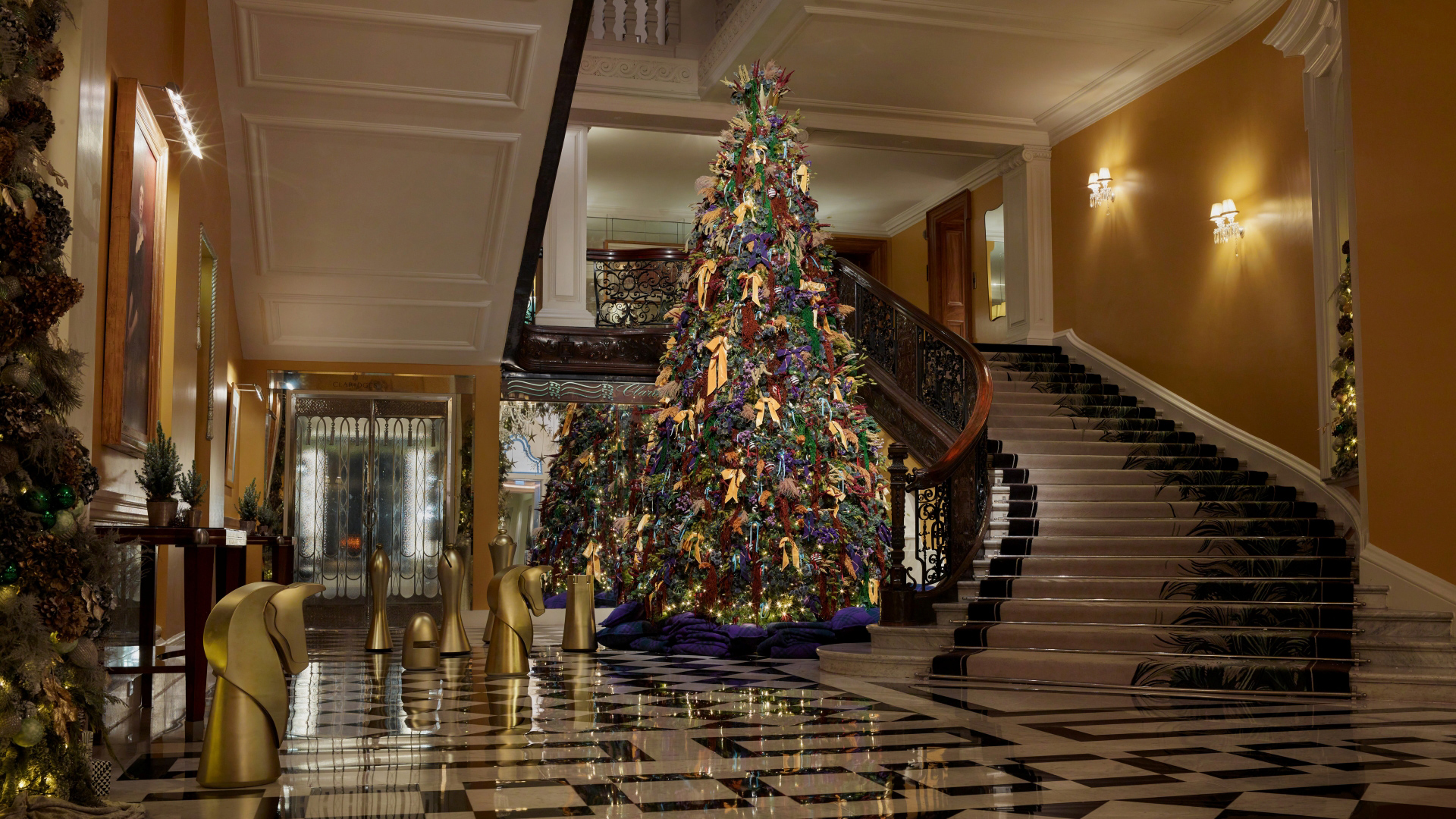 A photo of the annual Christmas tree in the Claridge's hotel foyer covered in fabric bows next to the grand staircase with giant black and gold chess pieces dotted around the floor in front
