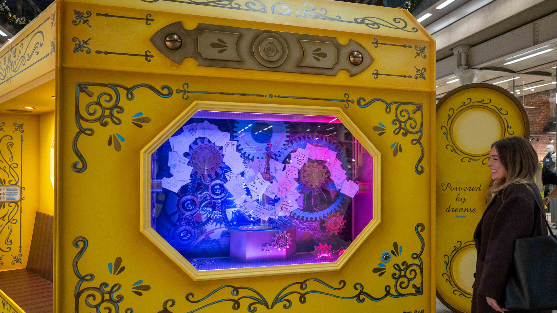 a close up photo of the bottom of the 2025 Christmas tree inside St Pancras International station with a giant yellow wind up music box filled with cogs and a women stood next to it smiling