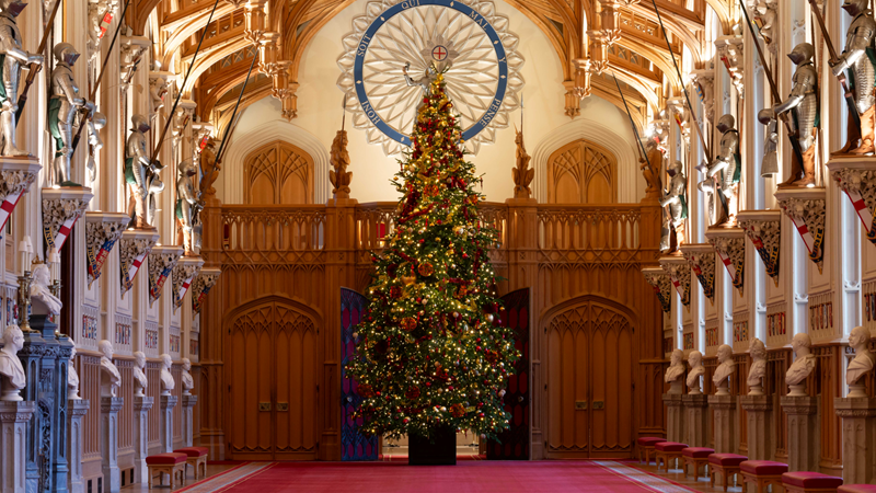 A large traditional Christmas tree with gold and red baubles stands in the centre of one of Windsor Castle's very grand rooms with red carpets, wooden features and armour hanging on the walls