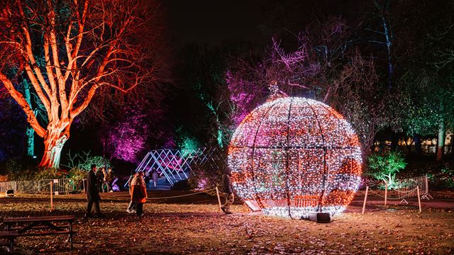 Visitors gathered around a large spherical Christmas decoration made of red twinkling lights, set in a dark outdoor area with trees illuminated in red and blue.