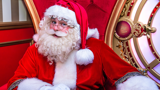 Santa Claus dressed in a traditional red and white suit sitting in a festive grotto at Hamleys, with a red throne and ornate gold detailing behind him.