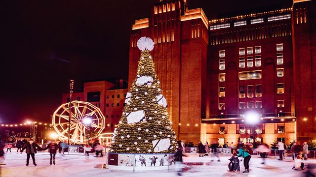 Un immense sapin de Noël illuminé se dresse au milieu d'une grande patinoire devant Battersea Power Station, où les gens patinent joyeusement avec des lumières de Noël en arrière-plan.