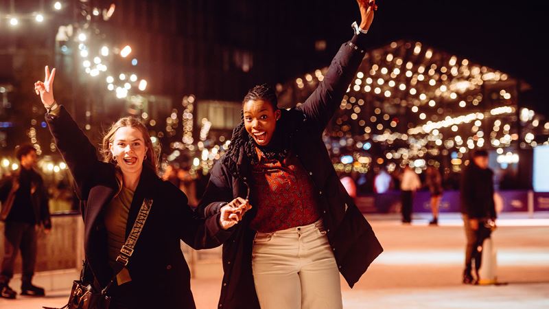 Deux femmes font du patin à glace à la patinoire Glide de Battersea, la nuit, profitant pleinement de leur séance et levant les bras en l'air.