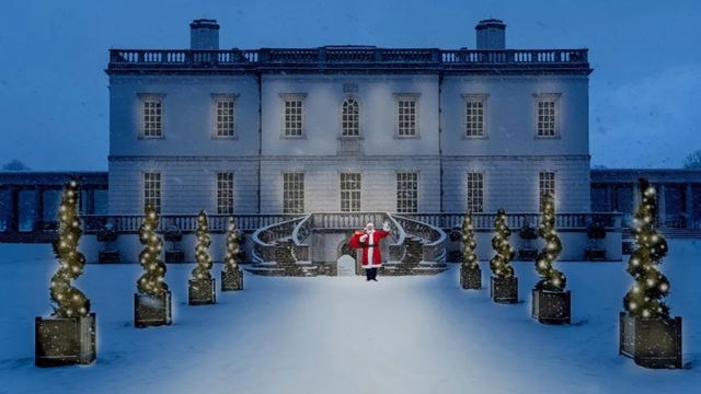 A large white mansion with pillars and windows and a tree lined walkway with festive lights and santa standing in the middle in a red suit and holding a black bag over his shoulder