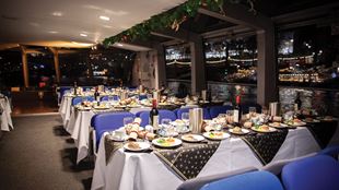 Ein heller Raum mit großen Fenstern auf einem Kreuzfahrtschiff auf der Themse mit grüner Weihnachtsdekoration und mehreren Tischen mit weißen und blauen Tischdecken, auf denen ein Weihnachtsbraten und Sektflaschen stehen.
