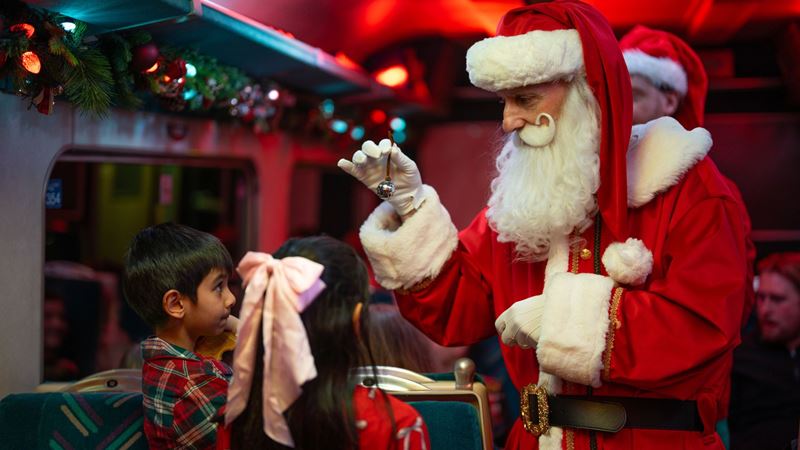 Santa Claus greets children inside a decorated train carriage during THE POLAR EXPRESS™ Train Ride, holding a silver bell as part of the interactive experience.