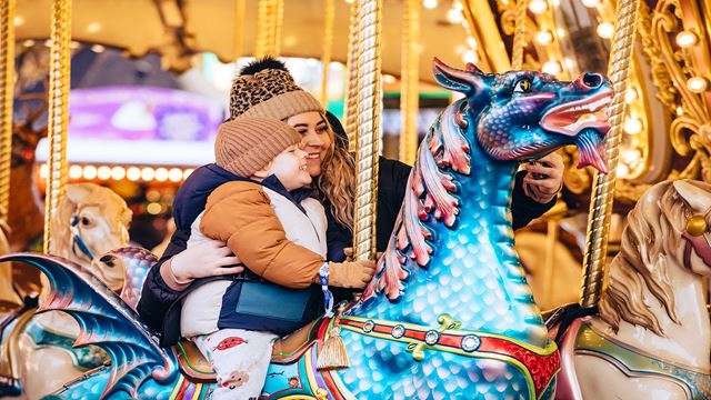 Woman accompanying young boy sitting on a blue dragon on a carousel ride.