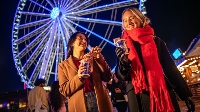 Zwei junge Frauen trinken heiße Schokolade im Winter Wonderland, im Hintergrund steht ein Riesenrad. 