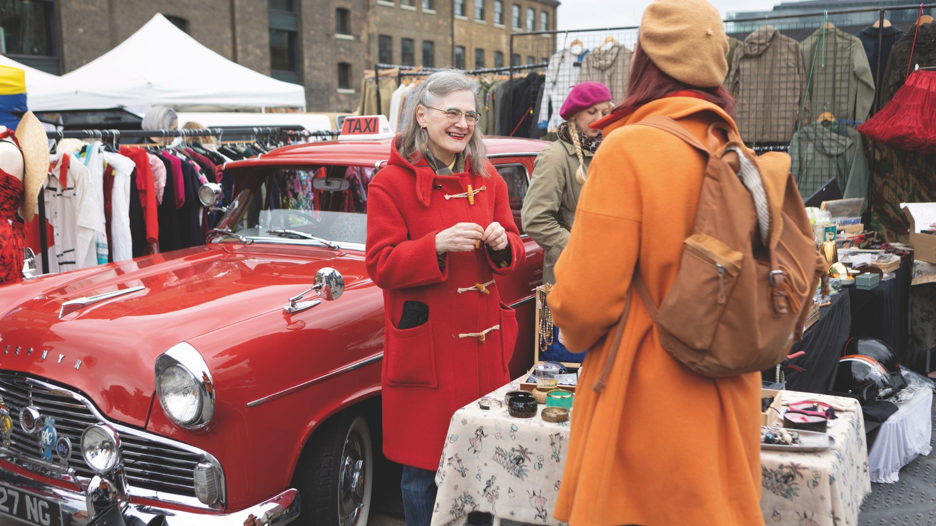 Enjoy classic cars and vintage shopping at the King's Cross Classic Car Boot Sale. Image courtesy of Classic Car Boot Sale - John Sturrock. People in brightly coloured coats chat at next to a red classic car, surrounded by stalls selling vintage clothes and items.