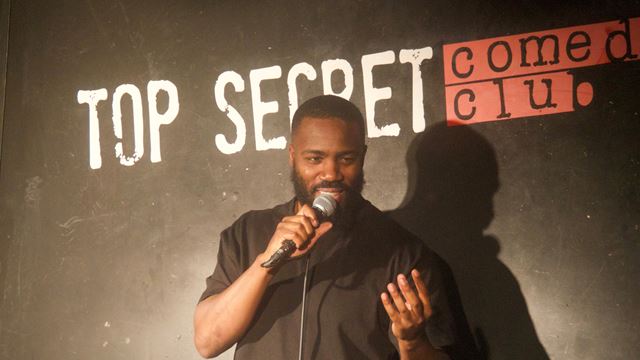 Mo Gilligan during a stand-up set, with the wording Top Secret Comedy Club on the set behind. 