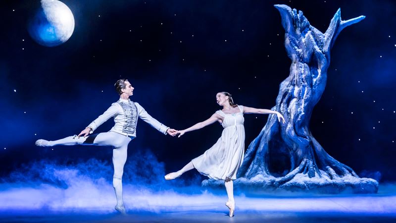 A male and female dancer wearing white, on stage, with a gnarly tree and large bauble in the background on the set of English National Ballet's Nutcracker.