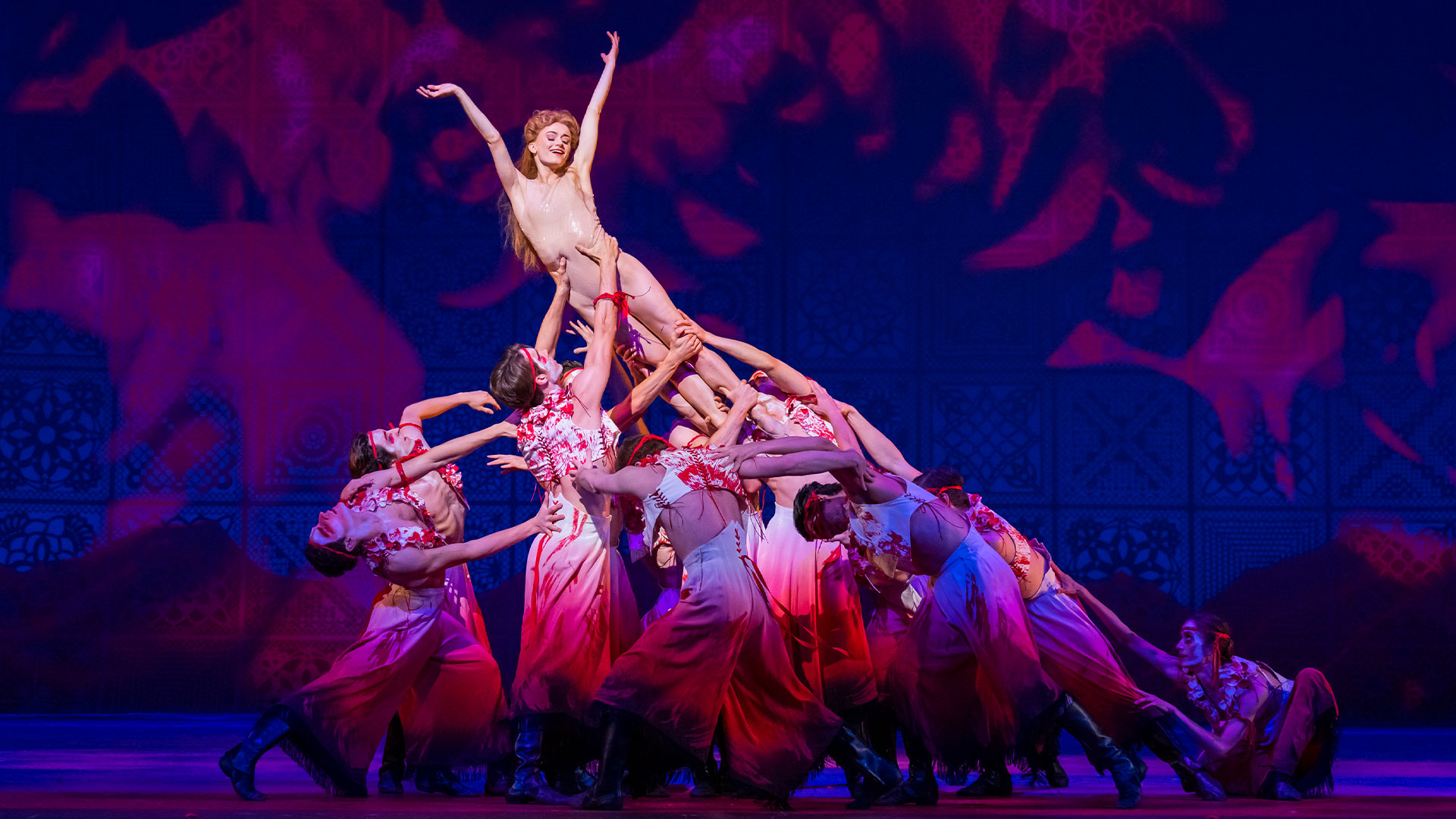 A ballerina wearing a sparkly beige leotard is lifted into the air by a group of ballet dancers wearing flowy red and white outfits.