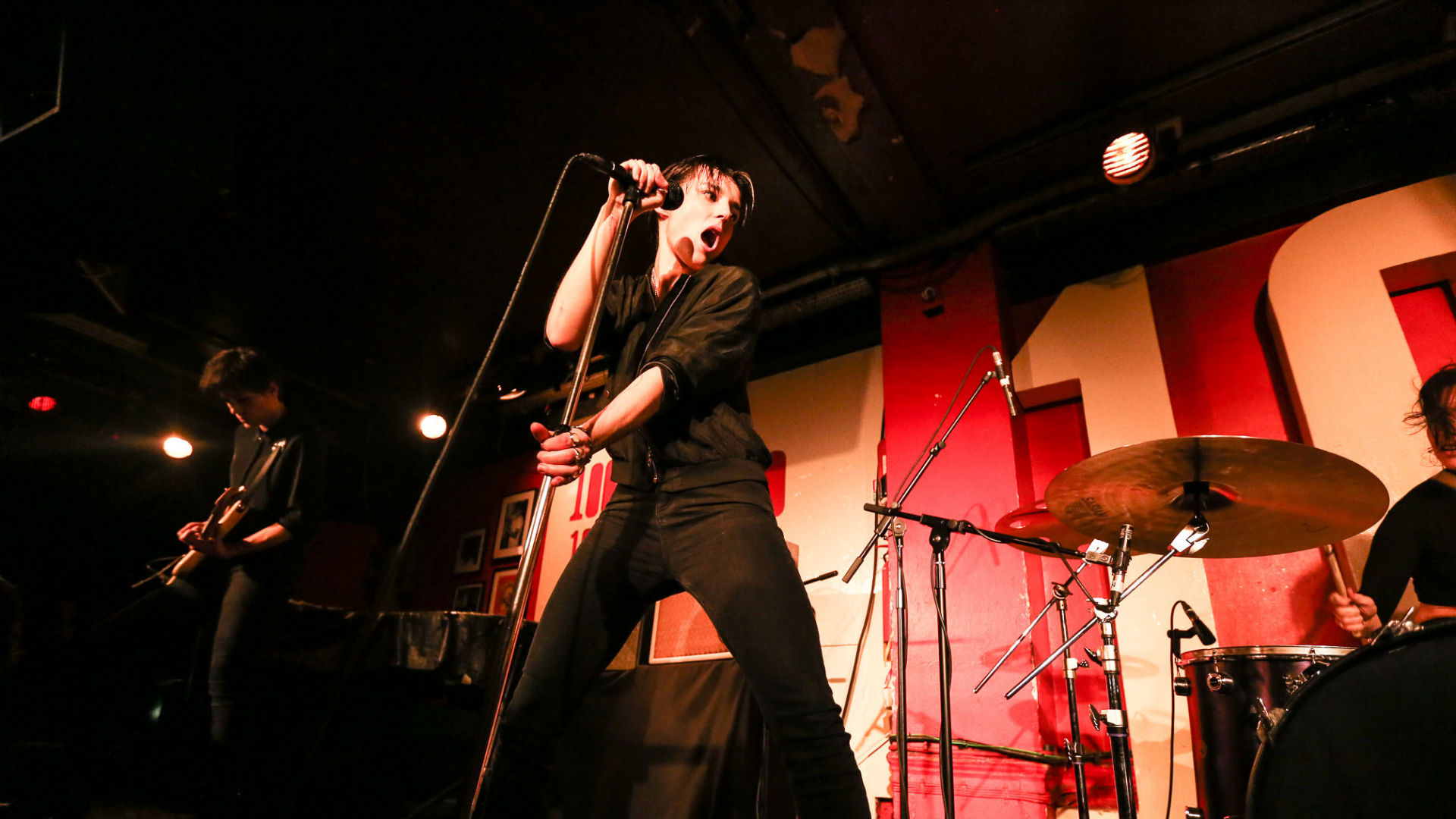 A person holding a standing microphone on stage, singing, with red background.