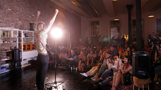 A photo of a person singing with their arms up as the audience watches, with a bright spotlight in the background.