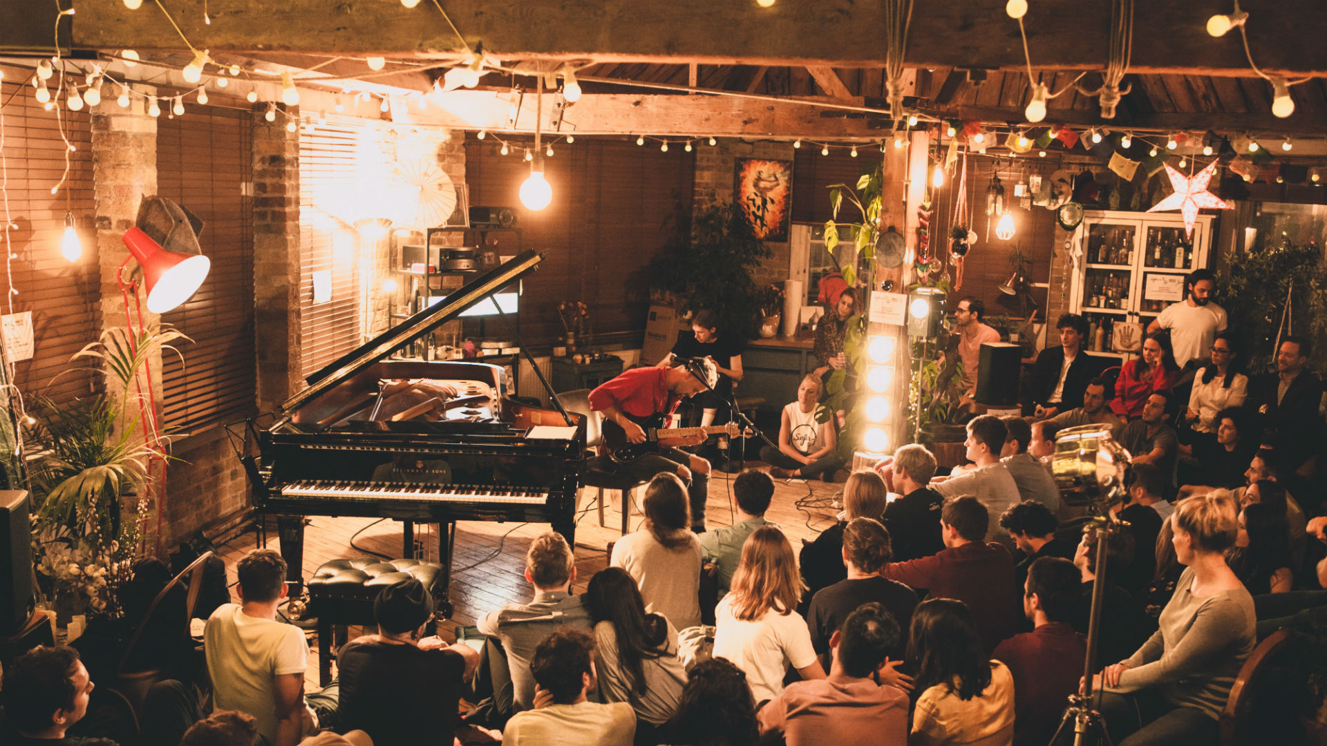 A photo of a man playing guitar, while the audience watches in a nicely lit room with a piano.