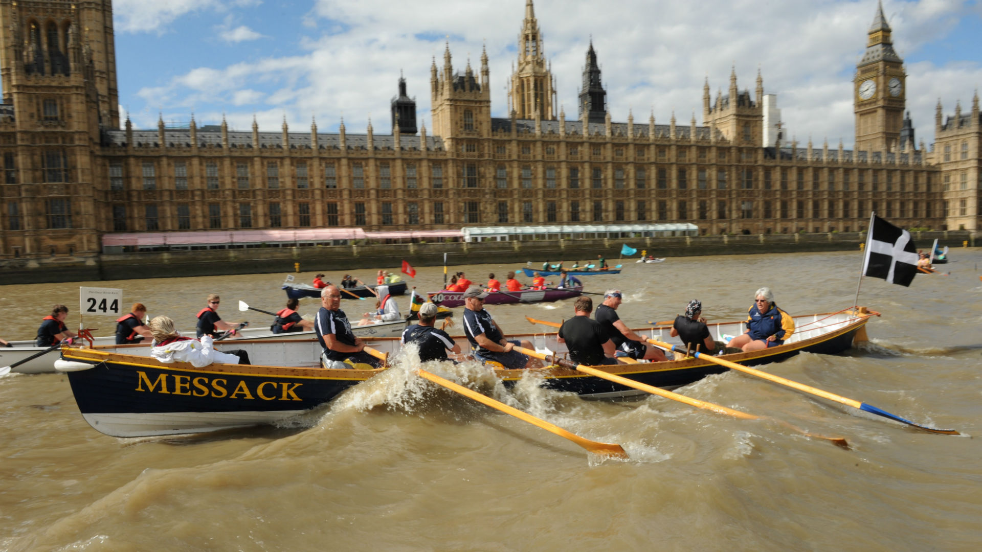 A photo of a rowing boat with people in it and the parliament buildings in the background. 
