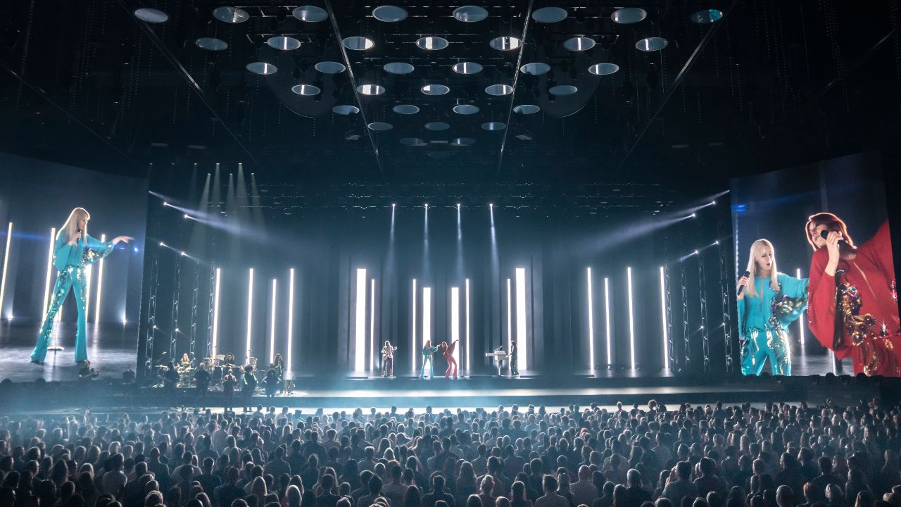 ABBA Voyage performance of Hole in Your Soul at London's ABBA Arena. Image courtesy of Johan Persson / ABBA Voyage. ABBA Voyage performers in green and red costumes, singing on stage in front of a large crowd at London's ABBA Arena.
