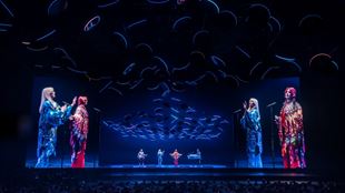 ABBA Voyage performers in blue and red costumes, singing on stage in front of a large crowd at London's ABBA Arena.