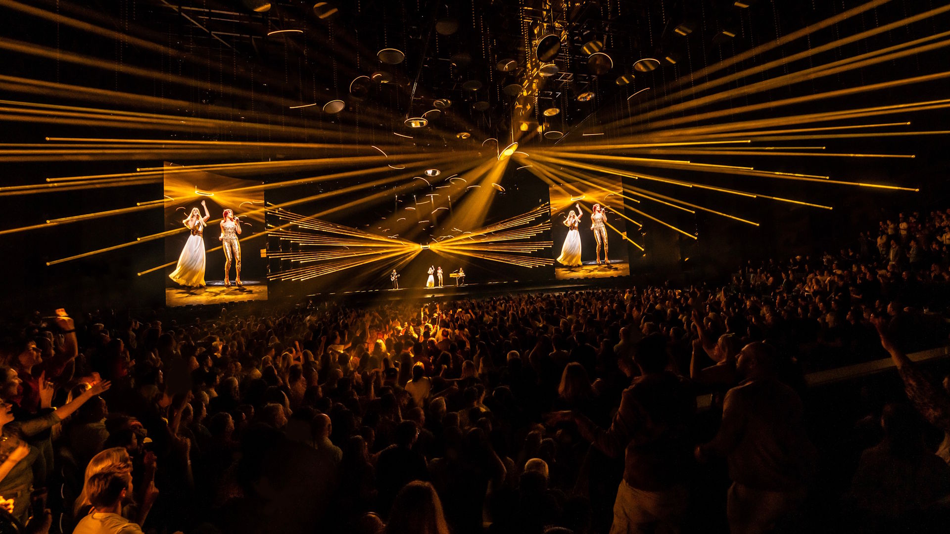 A crowd looks on in the dark as numerous yellow lasers shine out from the stage, with digital avatars of ABBA band members performing on stage.