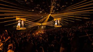 A crowd looks on in the dark as numerous yellow lasers shine out from the stage, with digital avatars of ABBA band members performing on stage.