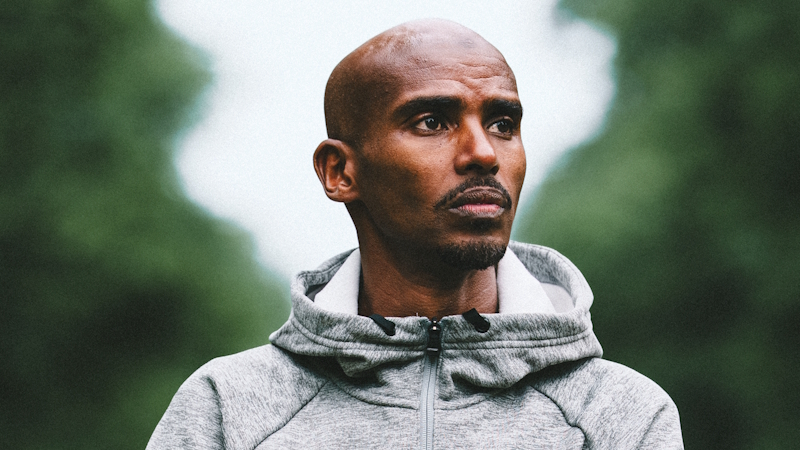 A close-up photograph of Mo Farah's head and shoulders, wearing a grey hoodie.
