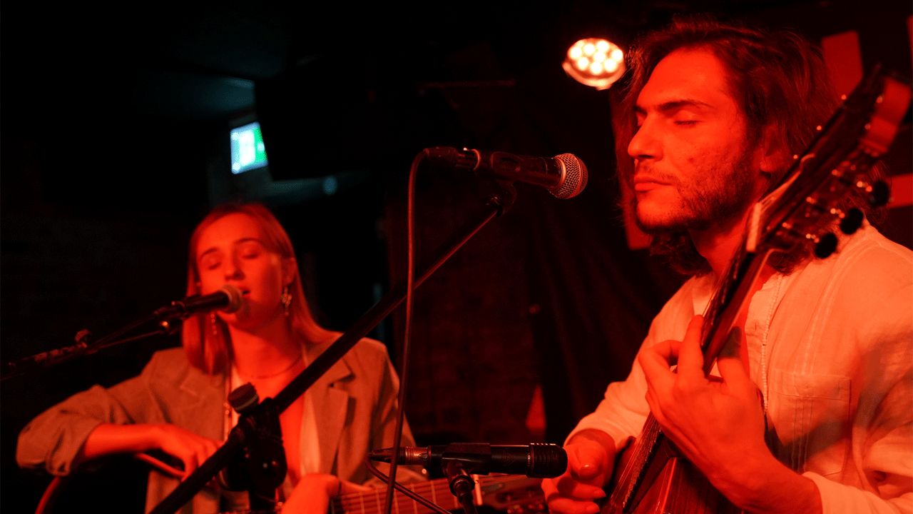 Man and woman sing whilst playing guitar illuminated by red lights at The Troubadour