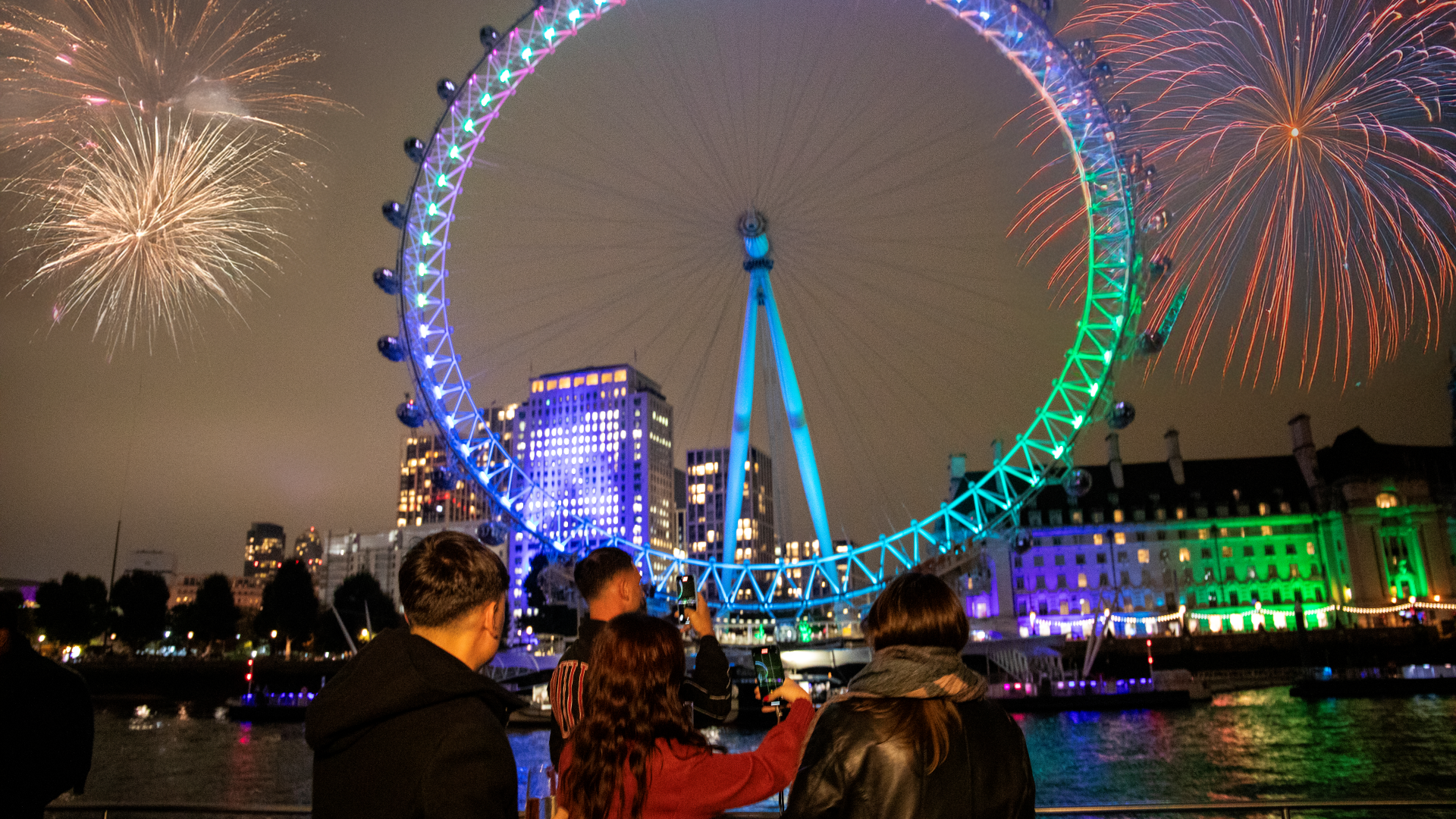 A group of friends watching the fireworks from a City Cruises boat as the London Eye lights up in purple and green next to the river Thames