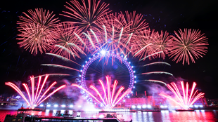 A photo of the London Eye with pink and blue lights surrounded by the London New Year's Eve fireworks display with pink fireworks in the sky, around the London Eye and coming from the river Thames