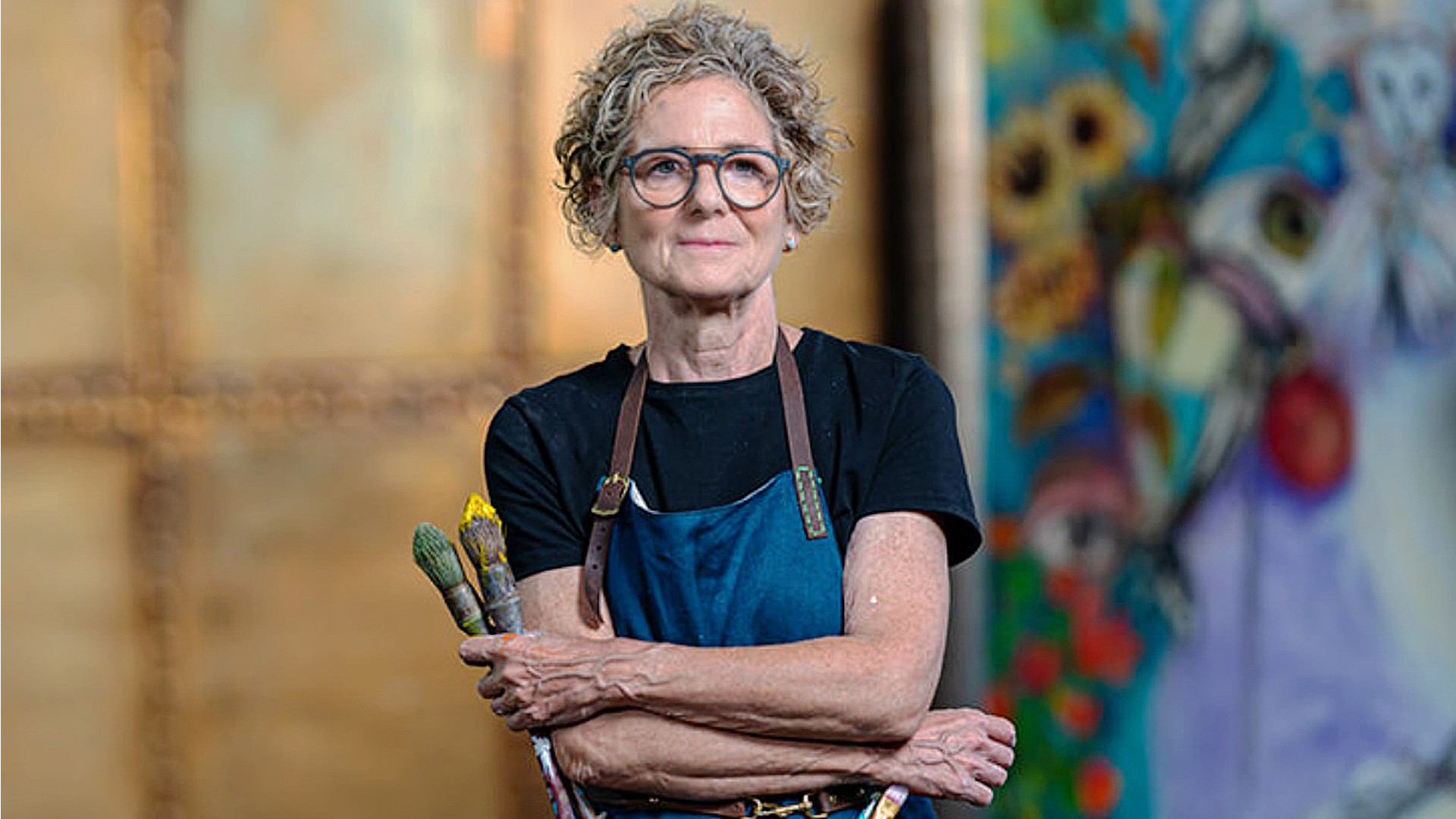 A portrait of artist Rachel Gadsden, holding paintbrushes, with an artwork in the background.