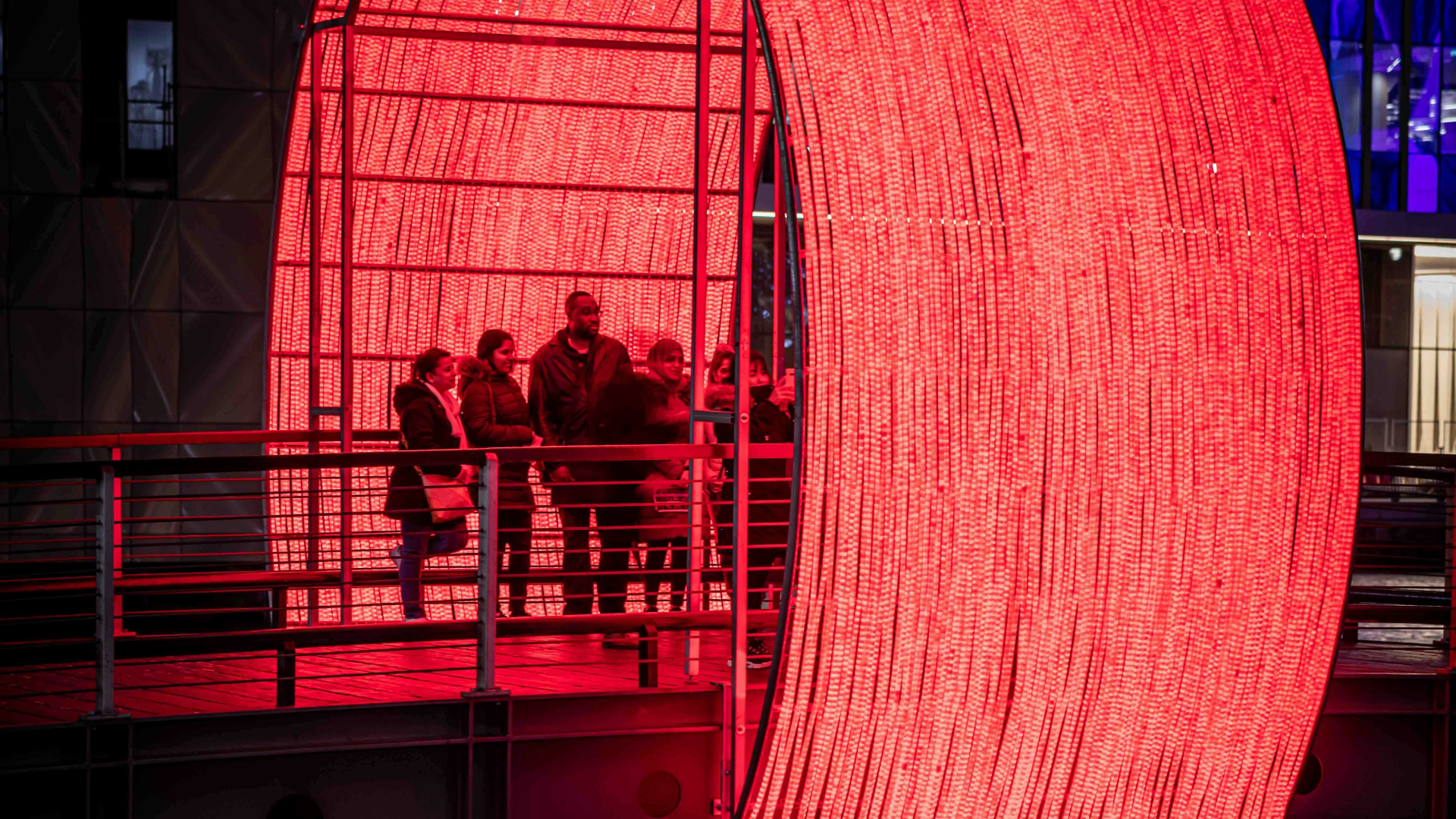 A group of people stood on a bridge at Canary Wharf with a huge glowing red light installation forming an archway around the bridge