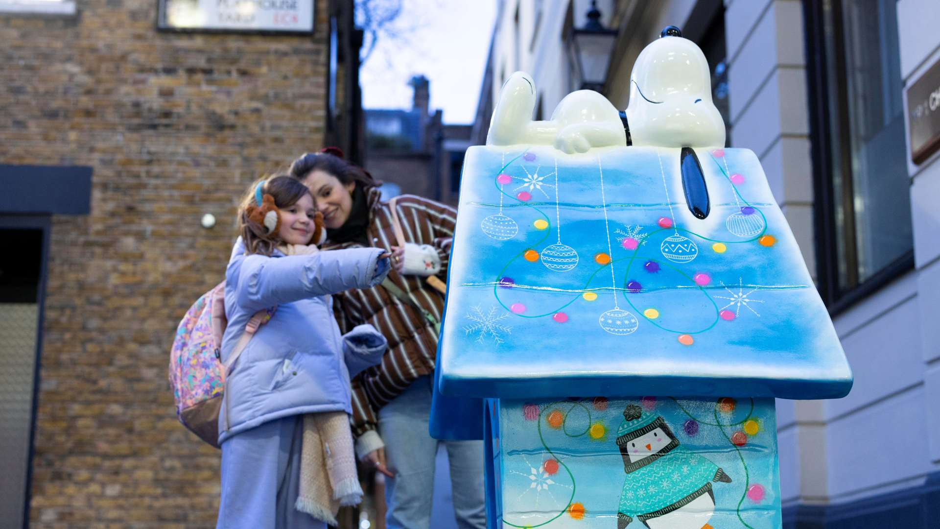 A photo of a mother with a young girl dressed in a purple coat with gloves and earmuffs pointing at a sculpture of Snoopy sleeping on his kennel along the Fleet Street Quarter trail