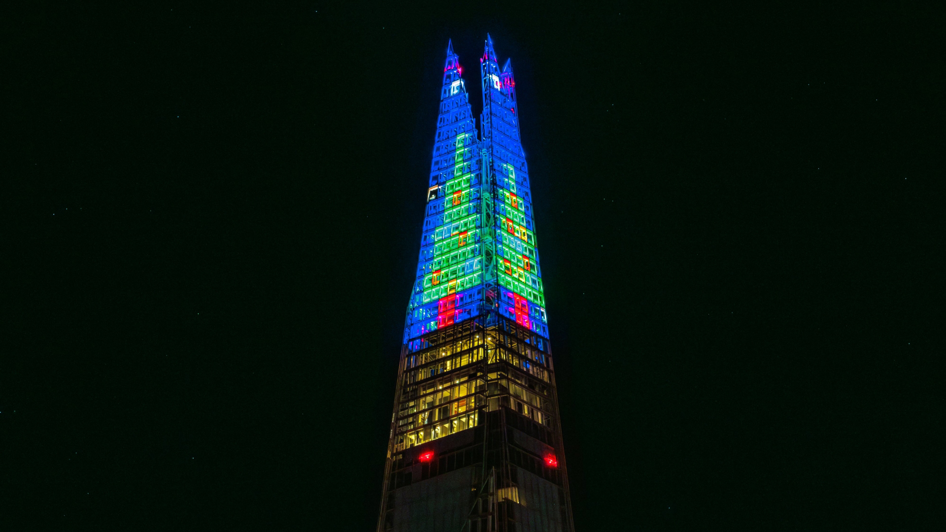 A photo of The Shard with a Christmas tree image made up of different coloured lights on the building's top floors, illuminating the sky