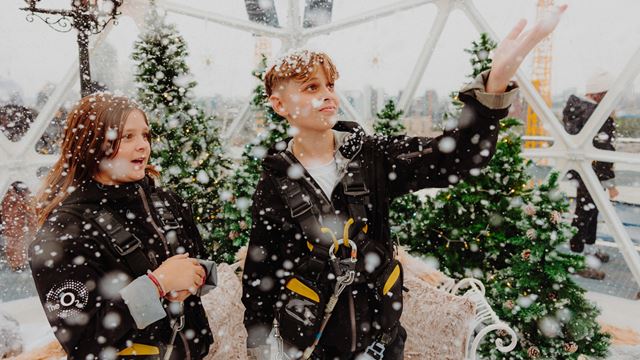 A photo of a young girl and boy stood inside a giant snow globe at the top of Up at The O2 with fake snow falling and Christmas trees in the background