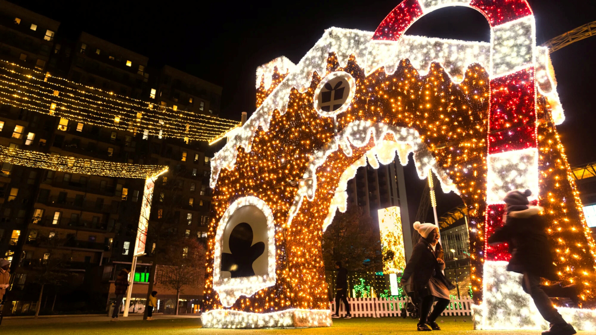 A photo of two children running around in front of a giant gingerbread light installation and candy cane at night