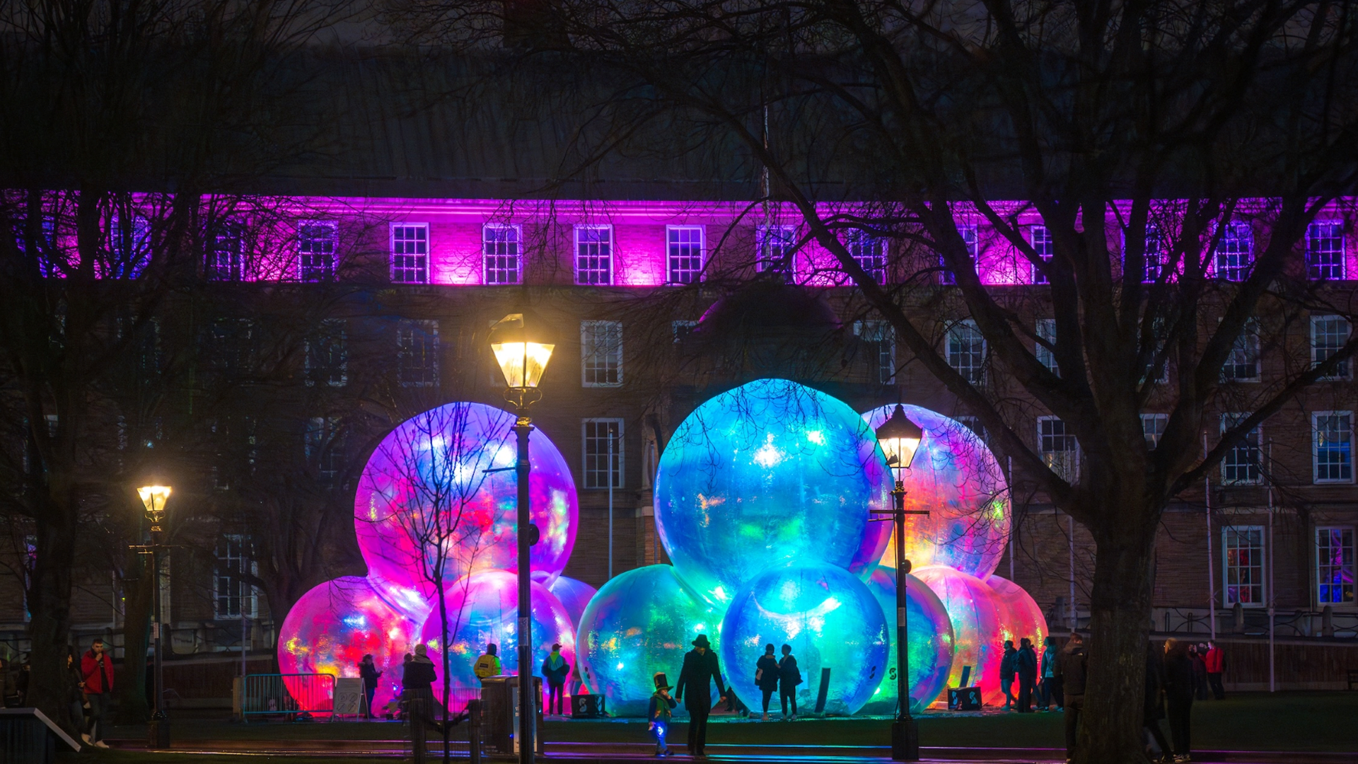 Step into a dreamlike world at Westfield London's Here We Glow, featuring light up installations from five different artists. Credit: Andre Pattenden. Image courtesy of Westfield London. A collection of large transparent bubble sculptures glowing shades of blue, pink and purple at night with outlines of people and lamposts in the foreground