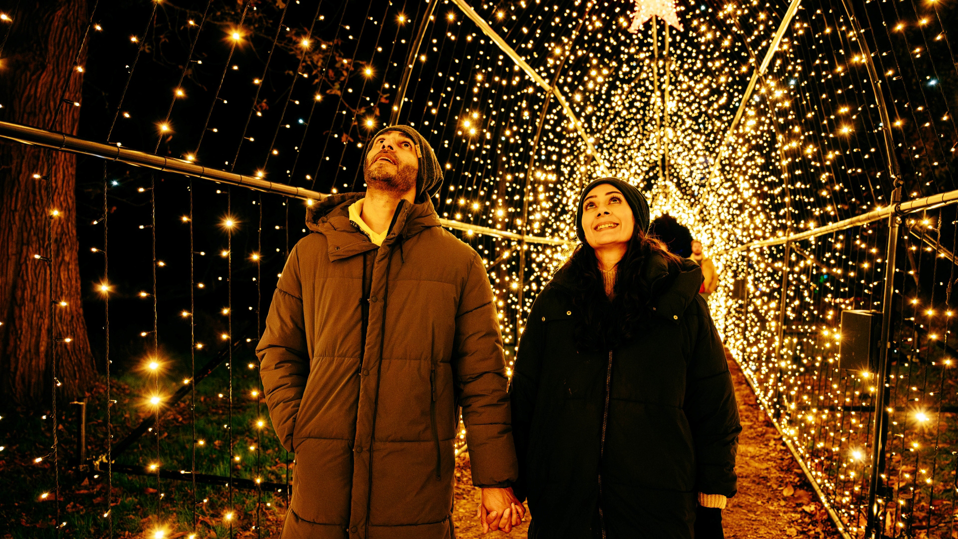 A photo of a man and woman dressed in thick coats holding hands inside a tunnel lined with twinkling lights