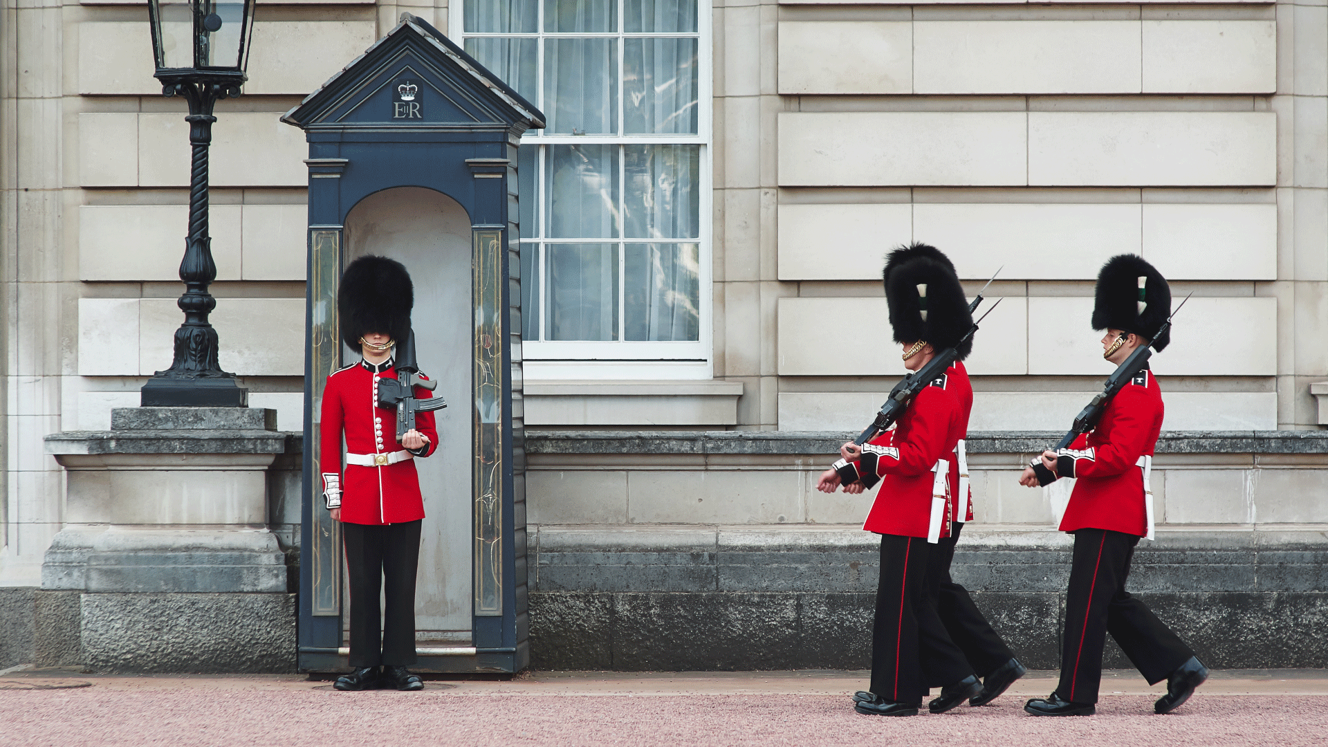 See guards dressed in traditional red tunics and bearskin hats during the Changing of the Guard. Image courtesy of Shutterstock. Two guards walk past another guard in traditional dress during Changing the Guard