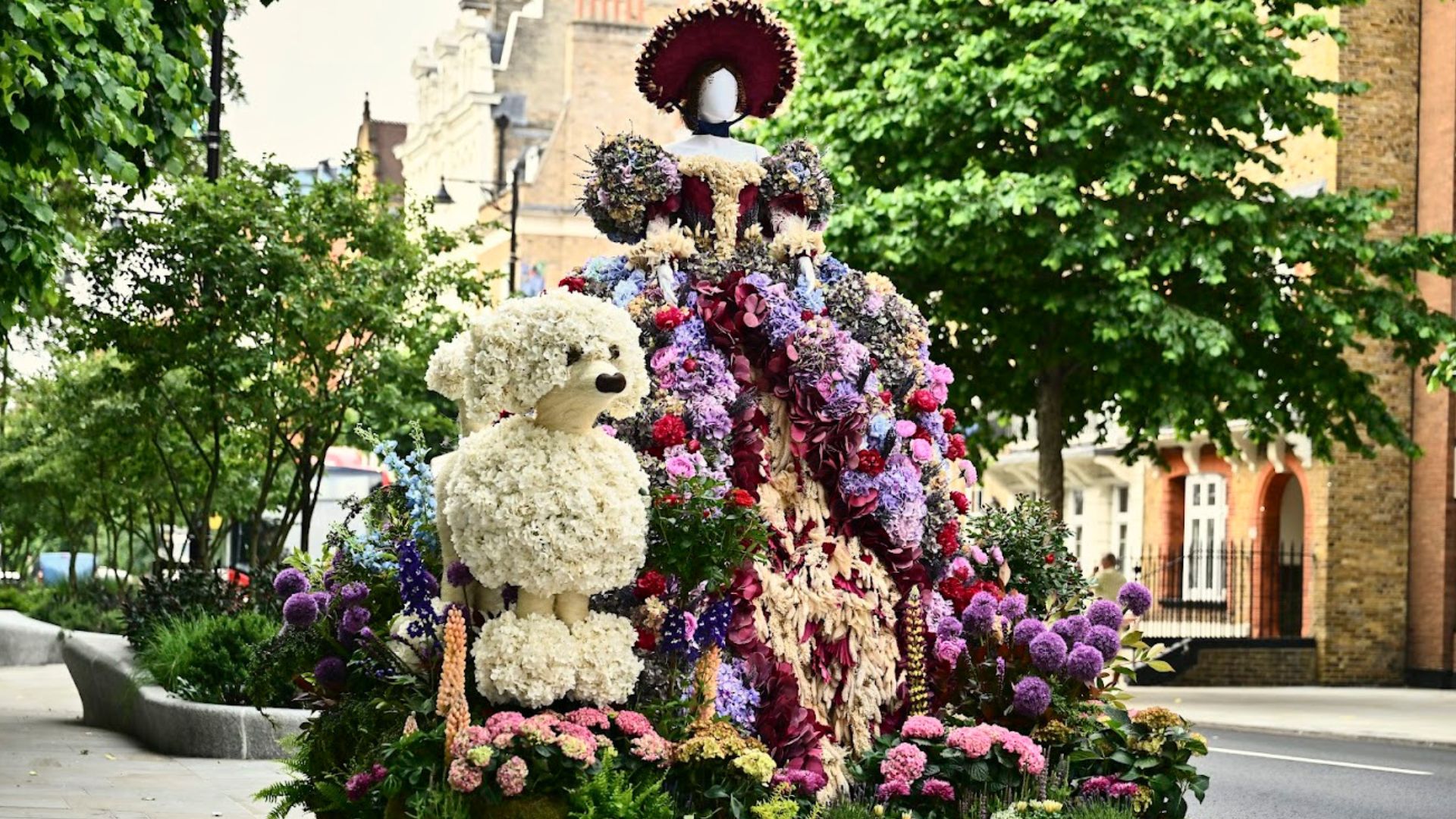A large free standing flower installation of a mannequin with a long flower flower dress and a poodle sat beside it also made from flowers